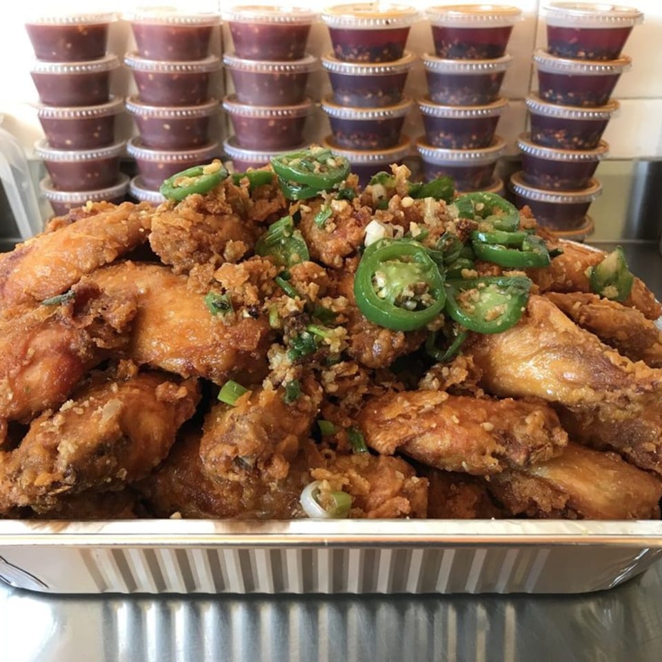 A large tray of crispy fried chicken wings topped with jalapeños and garlic, with sauce containers behind.
